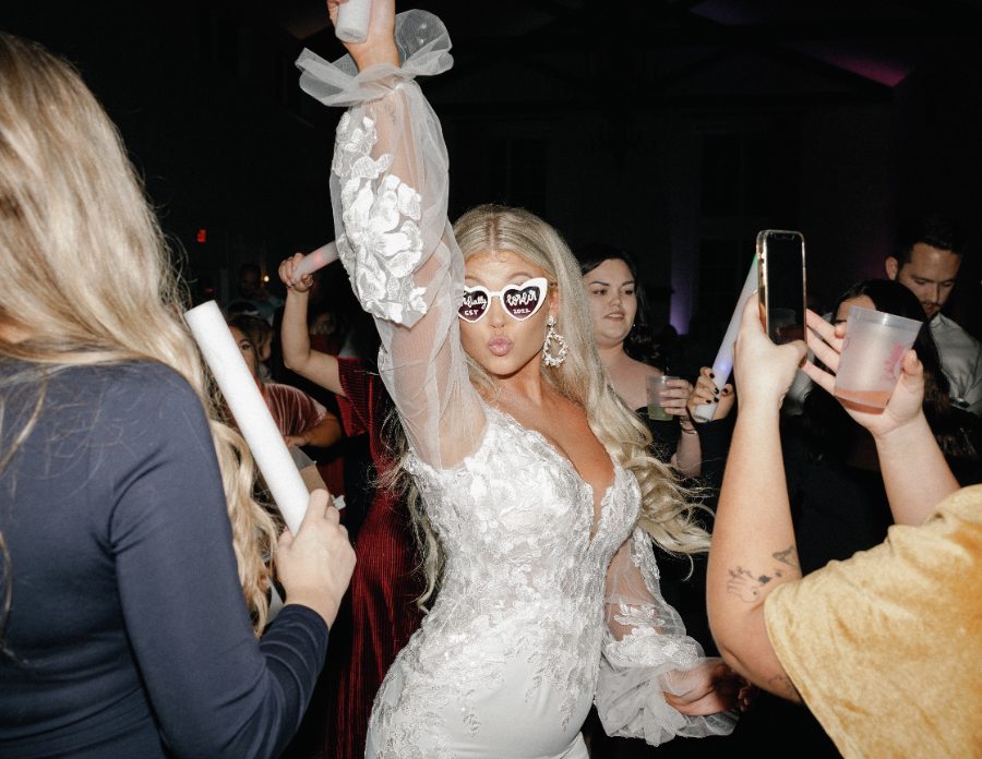 A bride in heart-shaped sunglasses dancing with a foam glow stick in the air
