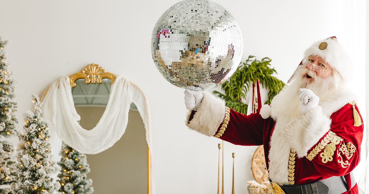 Santa in a red velvet suit balancing a mirrored disco ball on one finger, beside flocked Christmas trees