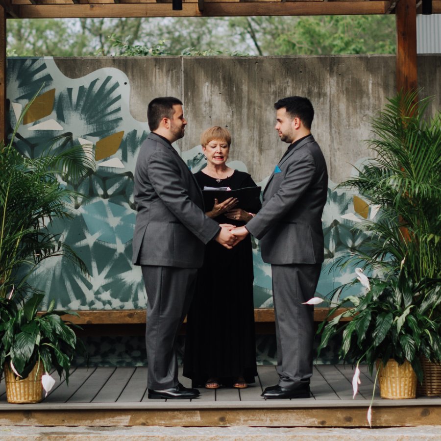 Two grooms holding hands during their ceremony under a wooden pergola