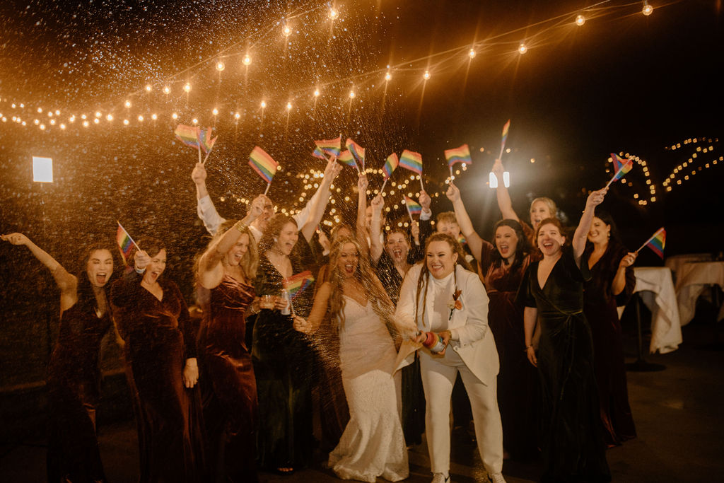 Two brides spraying champagne as their wedding party waves rainbow flags under string lights