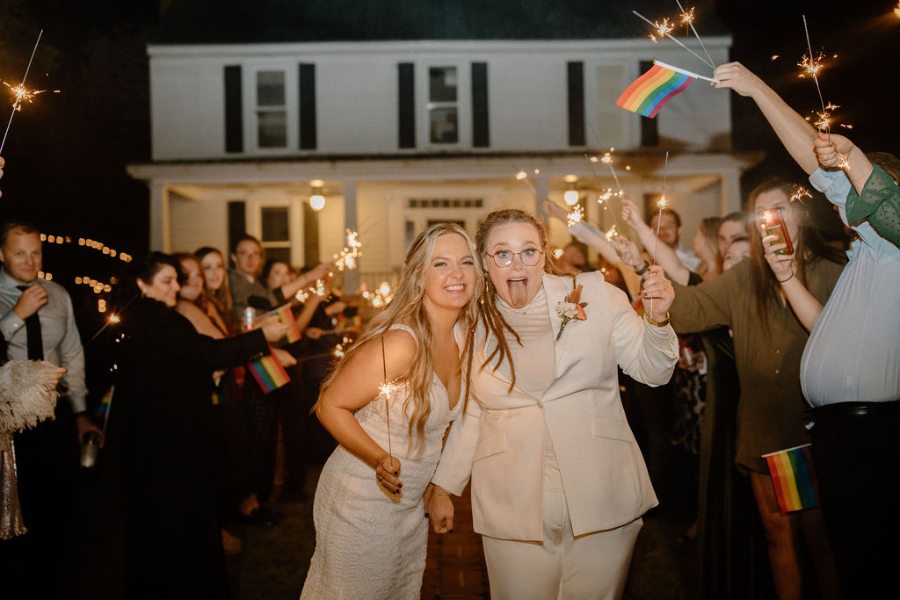 Two brides leaving through a sparkler send-off as guests wave rainbow flags
