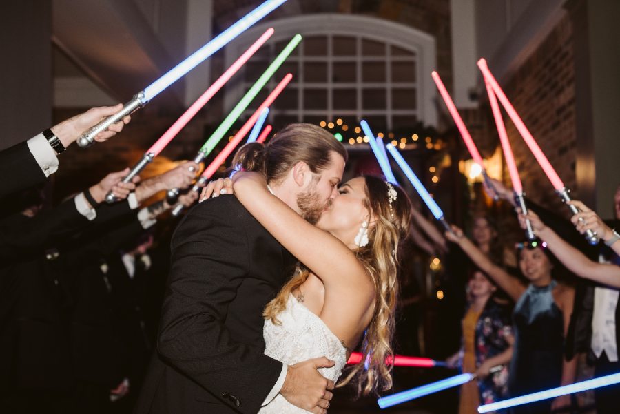 A couple kissing under a tunnel of light sabers held up by their guests at the end of the night